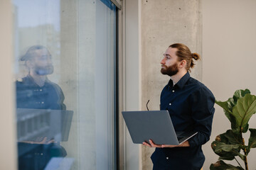 Young man holding laptop contemplating in modern office