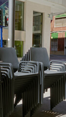 Stacked black chairs in sunlight cast shadows on city street with colorful buildings and pedestrians walking under a clear blue sky.