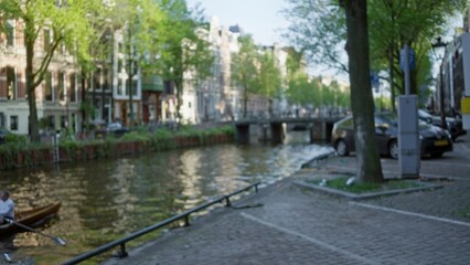 Urban canal scene in amsterdam with blurred buildings and a man rowing in a bokeh backdrop under the greenery of the netherlands.