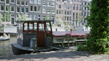 Blurred view of a houseboat docked along a picturesque canal in amsterdam with classic city architecture in the background and a focus on leisure and serenity.