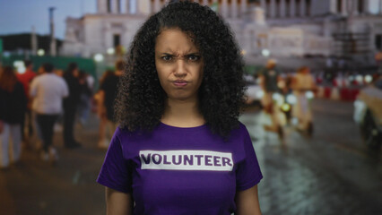 Young woman wearing a volunteer shirt stands on a bustling street at night, surrounded by people and city lights capturing an outdoor urban scene.