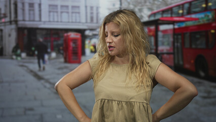 Woman, blonde, hands on hips, bare arms on a city street beside a red bus and telephone booth;...