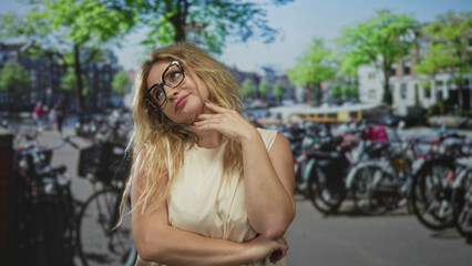Blonde woman with hand on chin and crossed arms at busy bike parking on a city street, wearing...