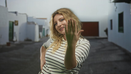 Woman beckons with hand and open palm on sunlit street between white buildings; playful invitation gesture.