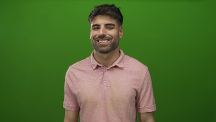 Fototapeta premium Young hispanic man in pink polo shirt smiles with slight head tilt in green studio setting; confidence.