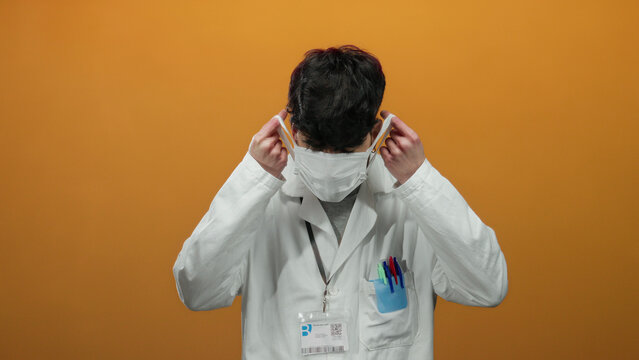Young man in a white lab coat adjusting face mask against a vibrant orange background, conveying a professional and cautious atmosphere.