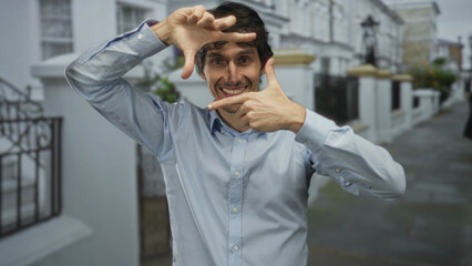 Obraz premium Smiling man framing with hands in front of white townhouses on city street; creativity imagination optimism vision.
