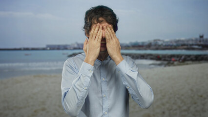 Man wearing light blue shirt covers eyes with hands in a gesture on a sandy beach near the sea;...