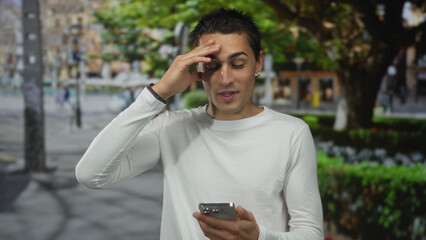 Young hispanic man wearing white shirt taps smartphone and touches forehead while looking aside on...