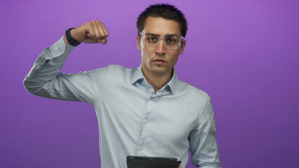 Young hispanic man holding a tablet confidently flexing his arm against a vibrant purple background exuding strength and modern technology prowess