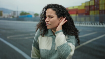 Fototapeta premium Woman at port making listening gesture, curly hair hispanic young adult against seaside with containers, wearing sweater and glasses, looks intently over sea during daytime.