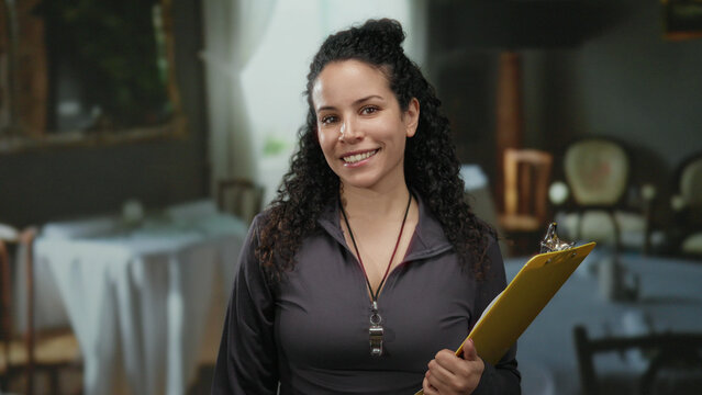 Woman smiling with clipboard in cozy restaurant setting showcasing a relaxed atmosphere with tables and chairs around her.