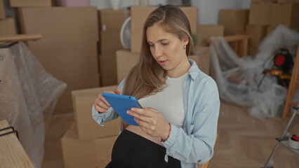 Pregnant woman sits in a new home living room with unpacked boxes, using a tablet, amidst an indoor moving scene implying a fresh start in her apartment.
