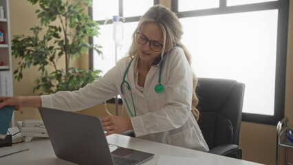 Woman doctor with stethoscope talking on phone using laptop in clinic room interior working.