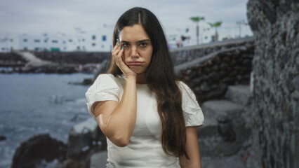 Woman in white shirt touching cheek on seaside by coastal building under cloudy sky with stone wall; boredom.