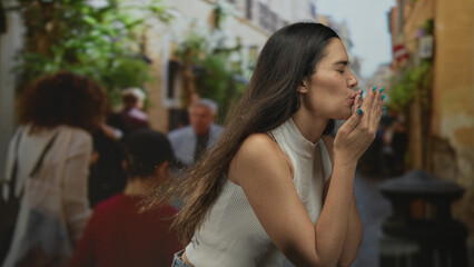 Young hispanic woman blowing kisses with hands to mouth while leaning forward in a narrow sunlit street; playful affection.