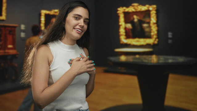Young hispanic woman clutching chest in gallery building with framed paintings on walls and wooden floor; gratitude.