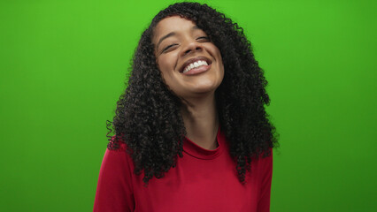 Woman smiles broadly showing teeth with curly hair and head tilt in green studio setting; joy...