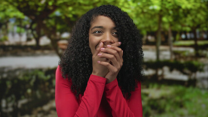 African american woman in long sleeved red shirt gently covers mouth with hands in sunlit forest;...
