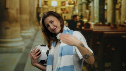 Woman smiling while holding a soccer ball and gesturing in a church setting, wearing a blue and white scarf, blend of sports and spirituality.