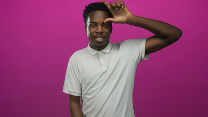 Man makes loser sign with hand on forehead while smiling in a bright pink studio; playful mockery...