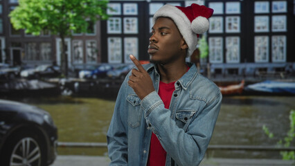 Man wearing red santa hat and denim jacket stands by canal on urban street looking sideways;...