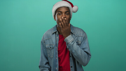 Young african american man in santa hat and denim jacket covering mouth with hand in teal studio; surprise.