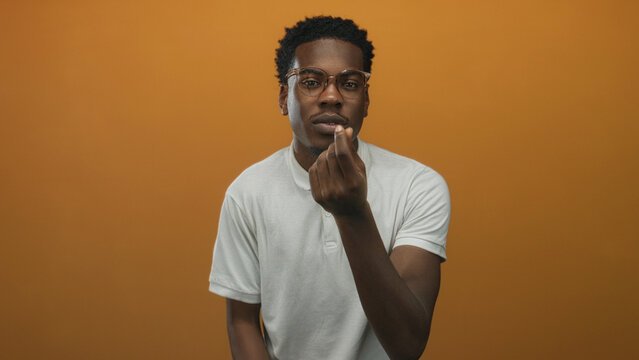 Young african american man rubbing his fingers in money gesture in orange studio setting for financial plea; desire wealth.
