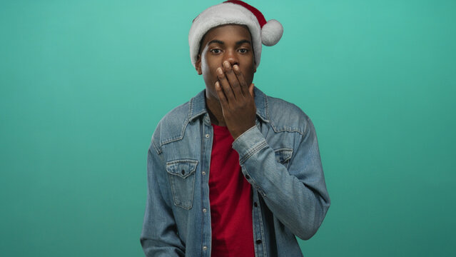 Young african american man in santa hat and denim jacket covering mouth with hand in teal studio; surprise. - Powered by Adobe