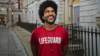Smiling man lifeguard wearing red shirt with text standing outdoors on city street, showcasing...