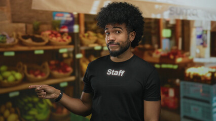 African american man in staff uniform points in an outdoor fruit shop with baskets of produce on shelves.