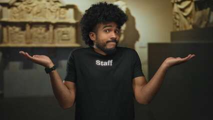 Man with afro shrugging while wearing staff uniform in museum indoor setting, standing against...