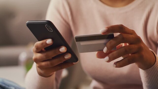 A young African American woman with curly hair sits comfortably at home, using her smartphone while holding a credit card, focused on making an online purchase.