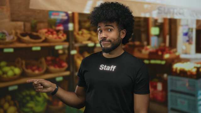 African american man in staff uniform points in an outdoor fruit shop with baskets of produce on shelves. - Powered by Adobe