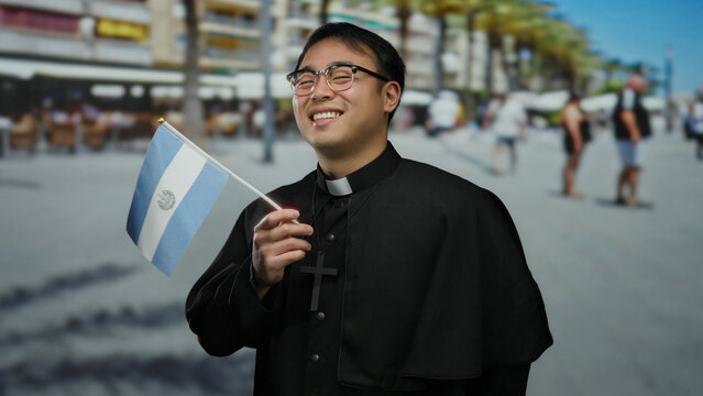 Asian man priest smiling holding salvadoran flag on busy urban street with buildings and people in background.