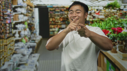 Young chinese man pointing finger amid flower pots in a building aisle lined with vibrant blooms;...