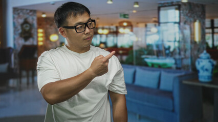 Young chinese man wearing glasses finger pointing to left in hotel lobby; uncertainty hesitation...