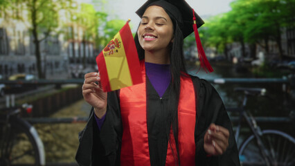 Smiling young latin woman in graduation gown and mortarboard holds spain flag on canal street; achievement pride.