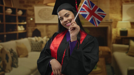 Smiling woman wearing graduation gown and mortarboard holding british flag in living room with sofa and shelves; achievement pride.