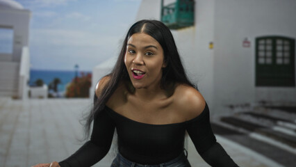 Woman smiles with open palms and bare shoulders against a white building facade on a seaside walkway; joy.