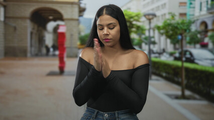 Latin woman hugging arms on street pavement lined with arches and lampposts in daylight;...