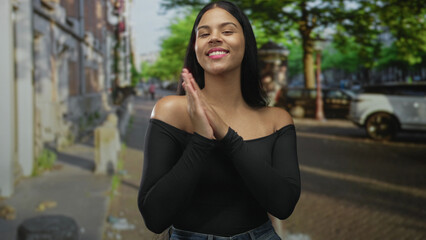 Woman rubbing hands together and smiling amid parked cars on a sunny city street in casual jeans...