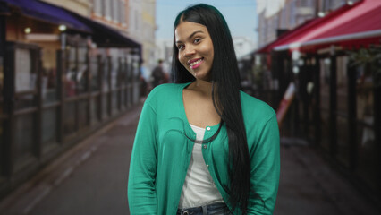 Latin woman with long dark hair in green cardigan smiles on city street outside cafe; confidence...
