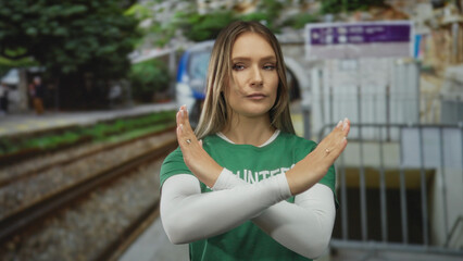 Woman volunteer in green shirt gestures rejection by crossing arms in an outdoor train station with blurred railway in the background.