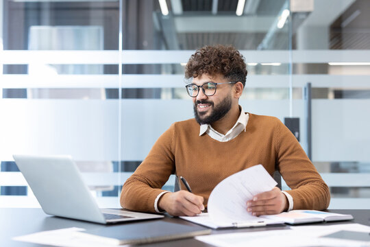 Young smiling businessman in glasses and casual wear sitting at desk, working on laptop and writing notes on documents, managing business tasks in a modern office environment - Powered by Adobe