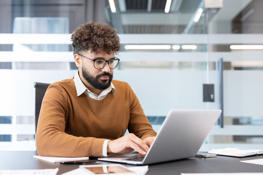 Focused young businessman with glasses and beard in a brown sweater working intently on a laptop at a modern office desk, typing and analyzing data for a project - Powered by Adobe
