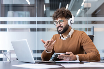 Smiling businessman with glasses and headphones leading a remote video conference from his modern office, using a laptop, taking notes and gesturing while discussing work and collaborating