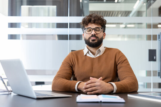 Young man meditating at his office desk, eyes closed and breathing deeply to relieve stress, promote mindfulness and calm during a busy workday in a modern workspace - Powered by Adobe