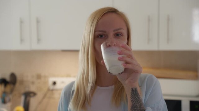 Happy blonde woman sipping on glass of refreshing milk in cozy kitchen, grinning at camera with playful milk mustache, relishing nutritious calcium-filled beverage