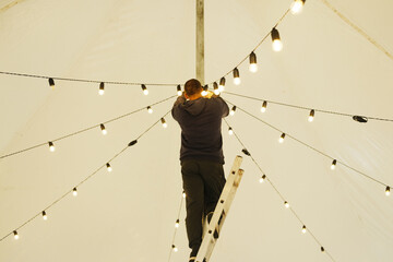 Male technician adjusting decorative string lights on a ladder inside a large tent, creating a warm ambiance for an outdoor event with illuminated atmosphere